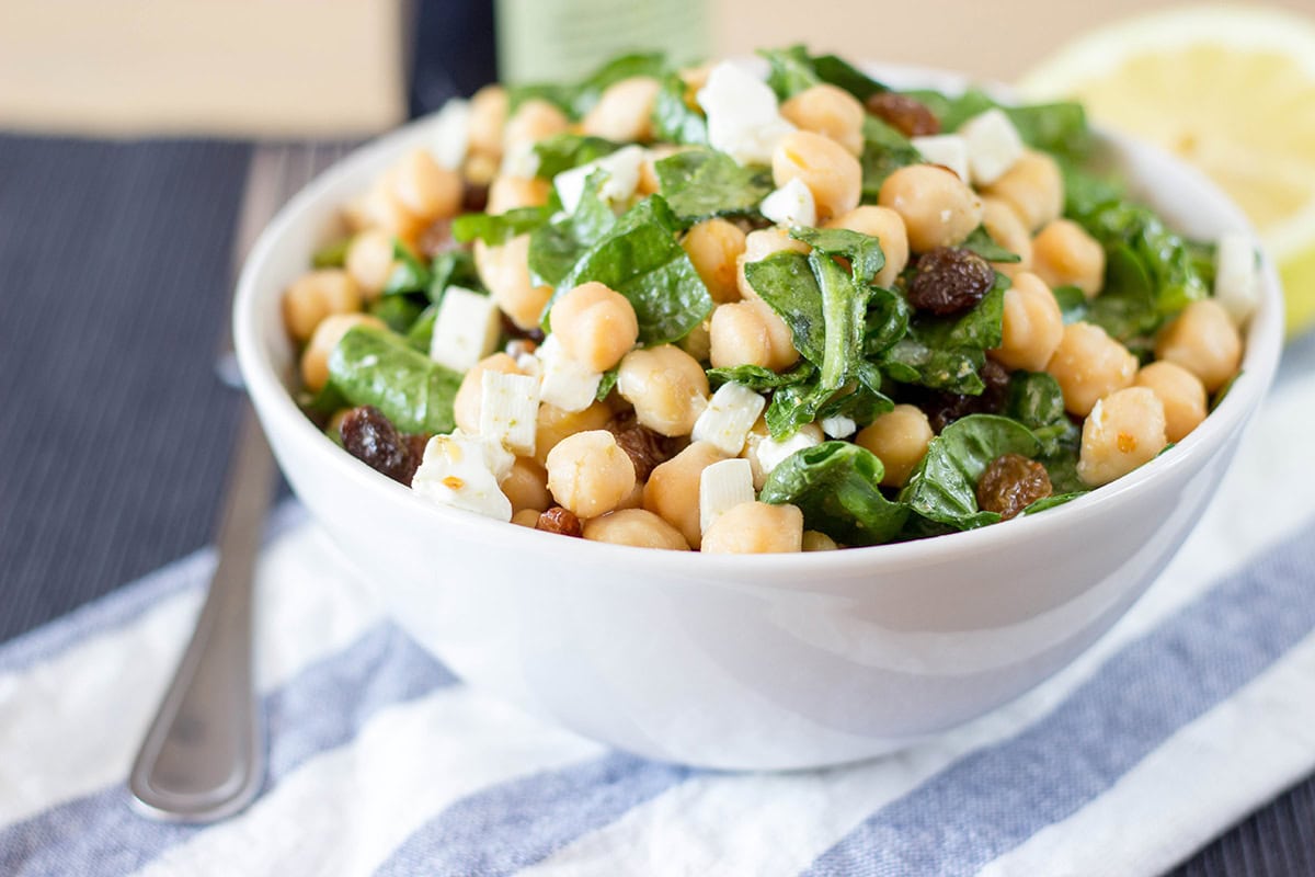 Chickpea Spinach Salad is served in a white bowl with a silver fork beside it, garnished with feta cheese and fresh herbs | Hurry The Food Up