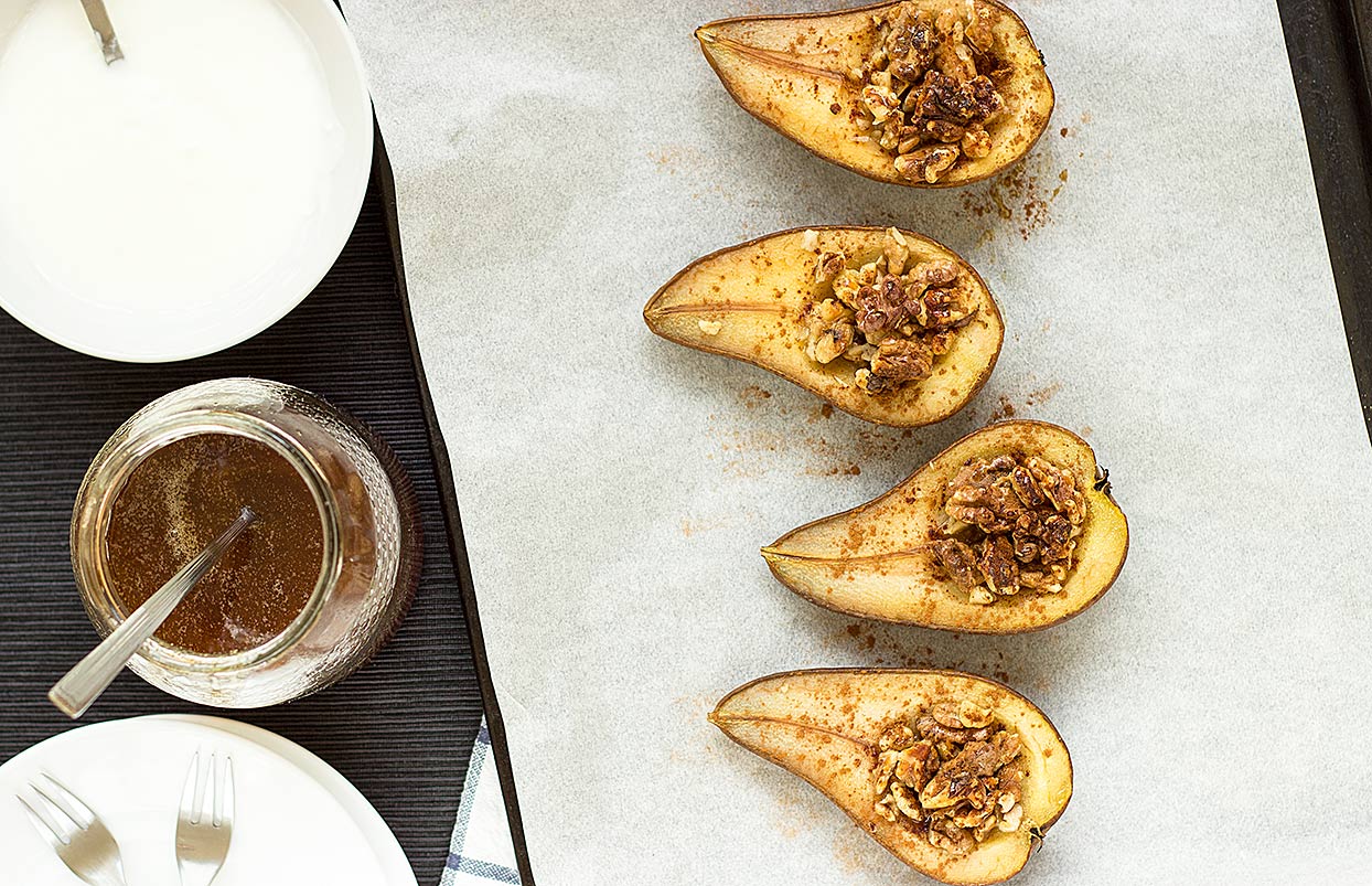 Bird's-eye view of four pear halves stuffed with walnuts and cinnamon. Next to them there are a jar of maple syrup with tea spoon, a bowl of soy yogurt and a plate with two forks | Hurry The Food Up