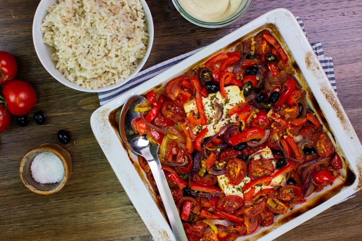 The dish is on the baking tray on a tea towel on a table with hummus, salt, tomatoes, olives and a deep plate with rice | Hurry The Food Up