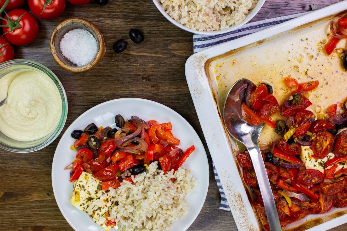 The feta veggie bake is served with rice on the plate on a table with hummus, salt, tomatoes, olives, and a bowl with rice | Hurry The Food Up