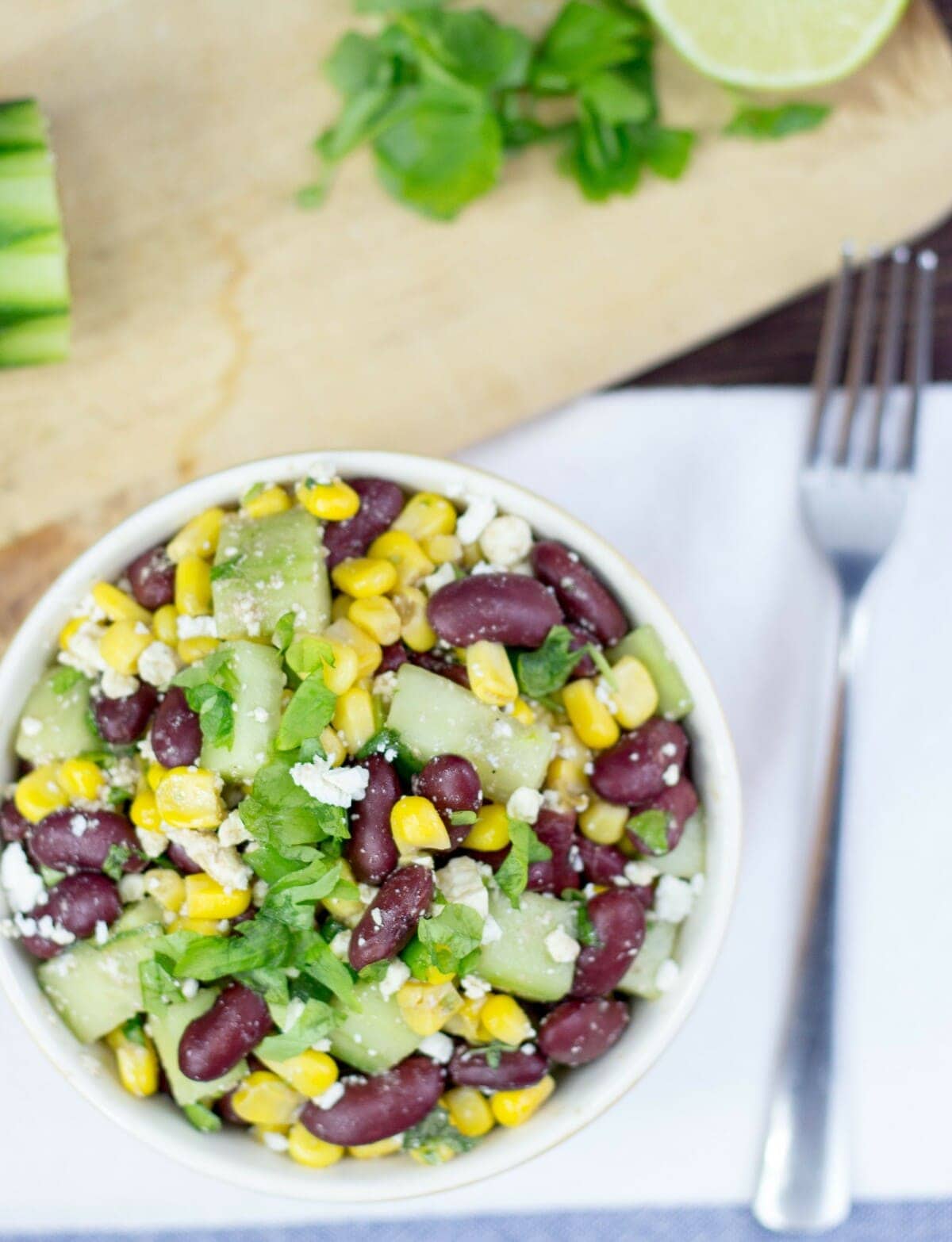 A bowl of high-protein kidney bean salad and a fork are placed over a white napkin. Parsley leaves and half a lime are on a wooden platter in the distance | Hurry The Food Up