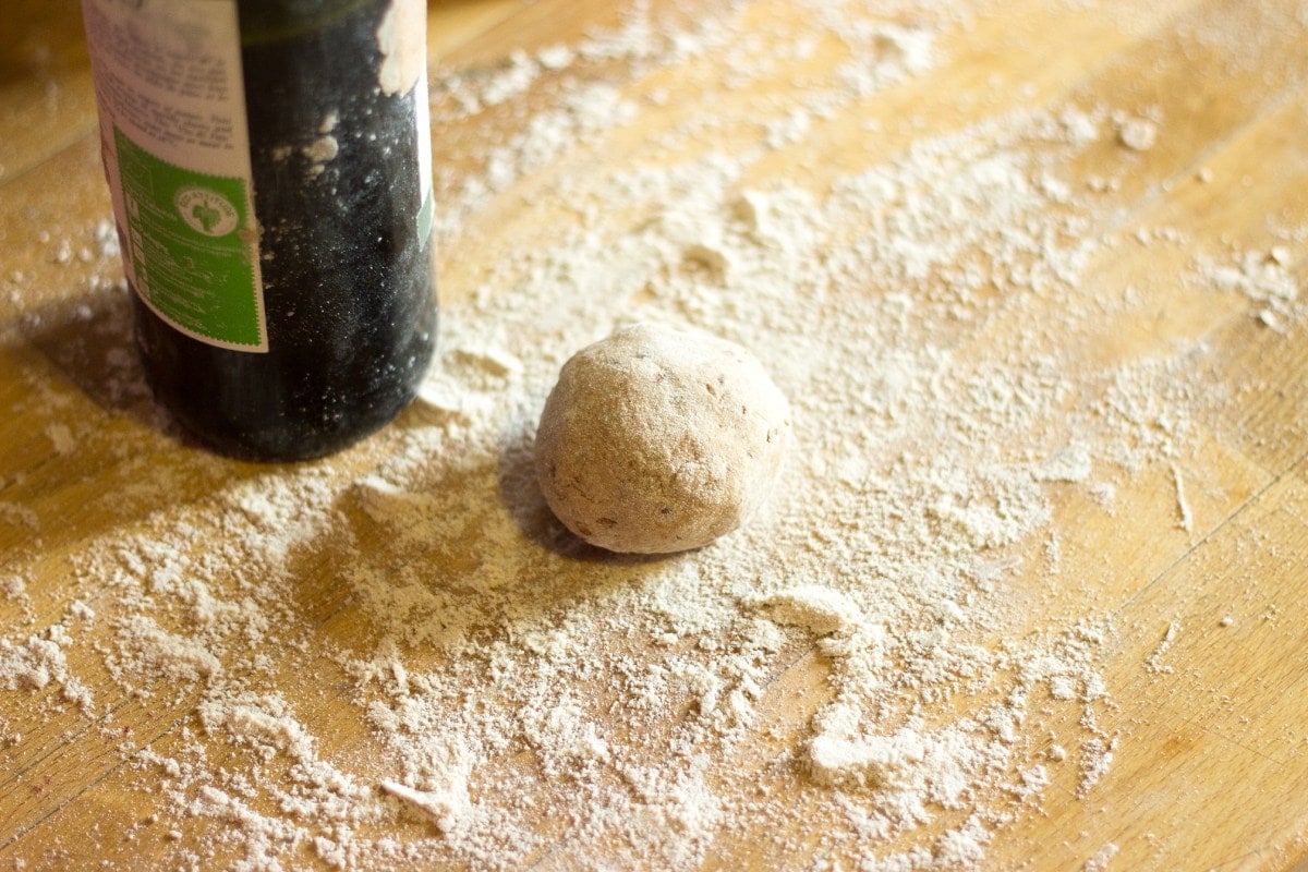 A piece of dough lies on the wooden surface sprinkled with flour. Next to it, there is a bottle of olive oil | Hurry The Food Up