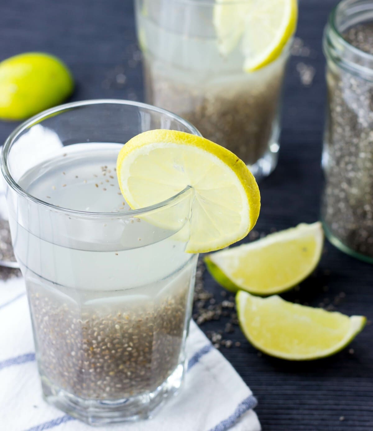 A glass of chia fresca (iskiate) placed on a white towel is seen with a slice of lime on its rim. Two other glasses and lime slices are in the distance | Hurry The Food Up