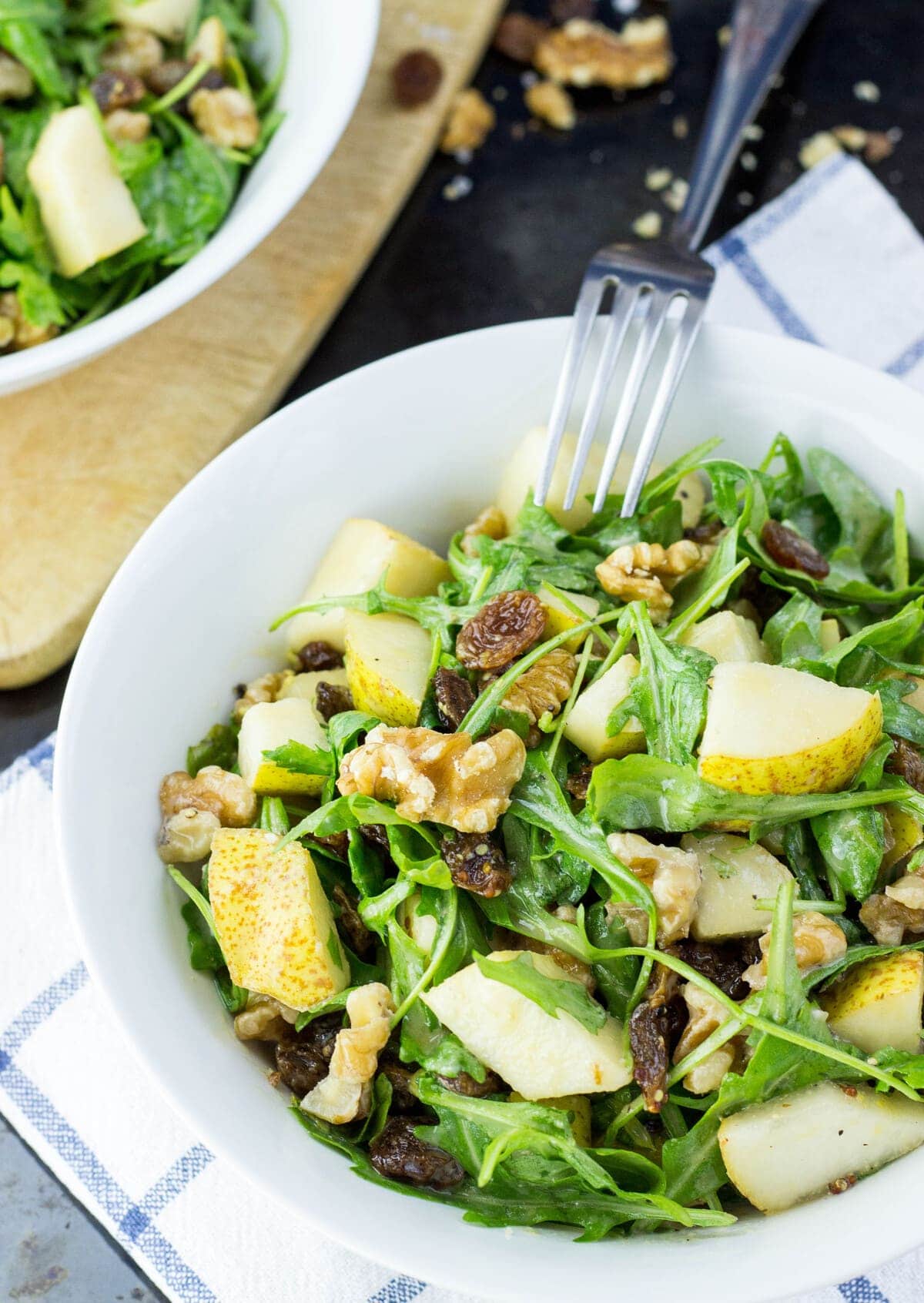 Pear and walnut salad is placed in a white bowl over a striped cloth with a silver fork. Another bowl of pear and walnut salad is in the distance. | Hurry The Food Up