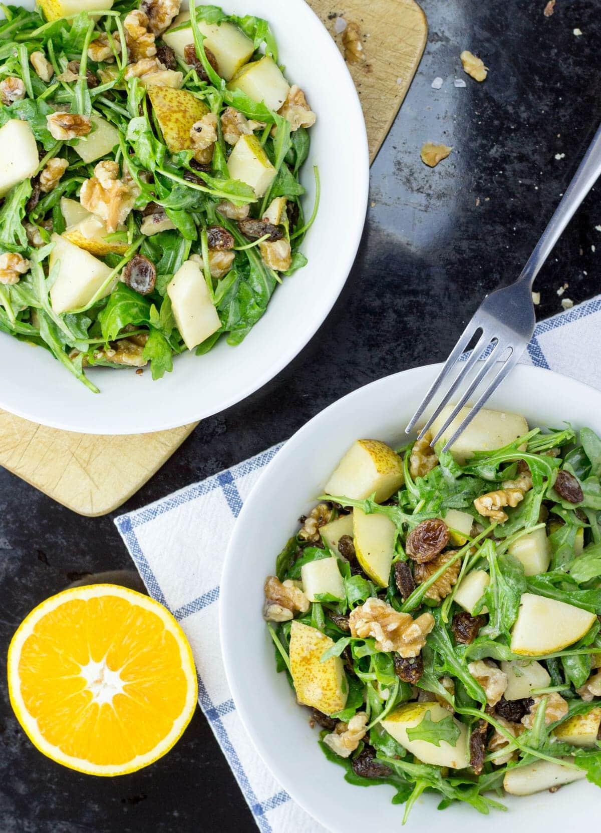 Two white bowls of pear and walnut salad are placed on a cutting board and cloth over a table with an orange and crumbs of walnuts | Hurry The Food Up