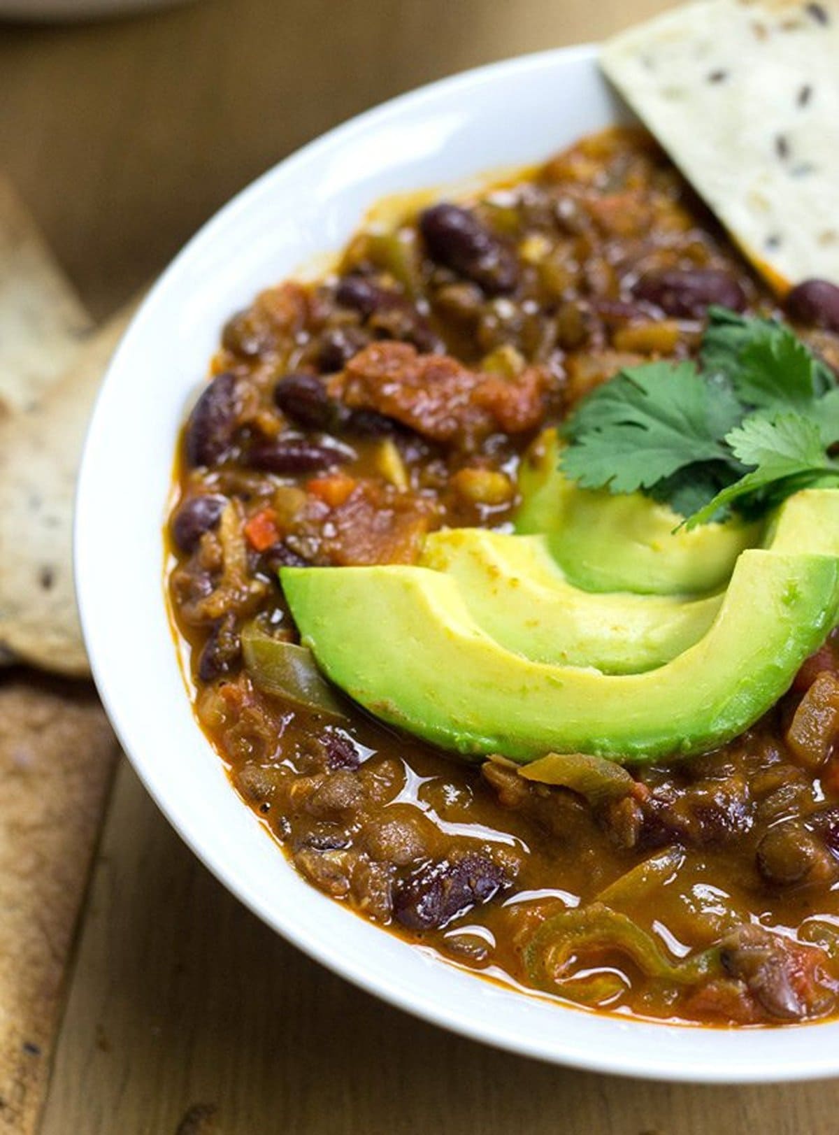 A close up of a white bowl filled with vegan chilli topped with avocado, sat on a wooden table | Hurry The Food Up