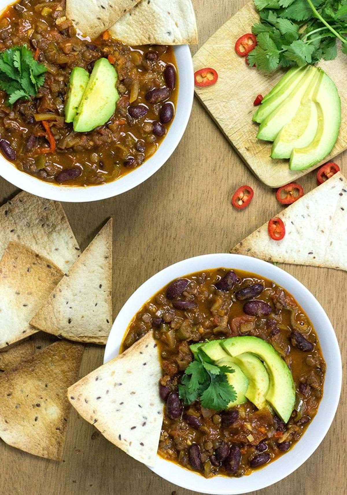 A birds-eye view of a wooden table. On the table are two white bowls of chili topped with avocado, next to them is a chopping board with more avocado, cilantro and chilis as well as toasted bread | Hurry The Food Up