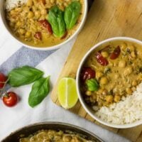 Two bowls with Vegan Chickpea Curry. Next to them, there are some tomatoes, fresh basilic leaves, and a piece of lime | HurryTheFoodUp