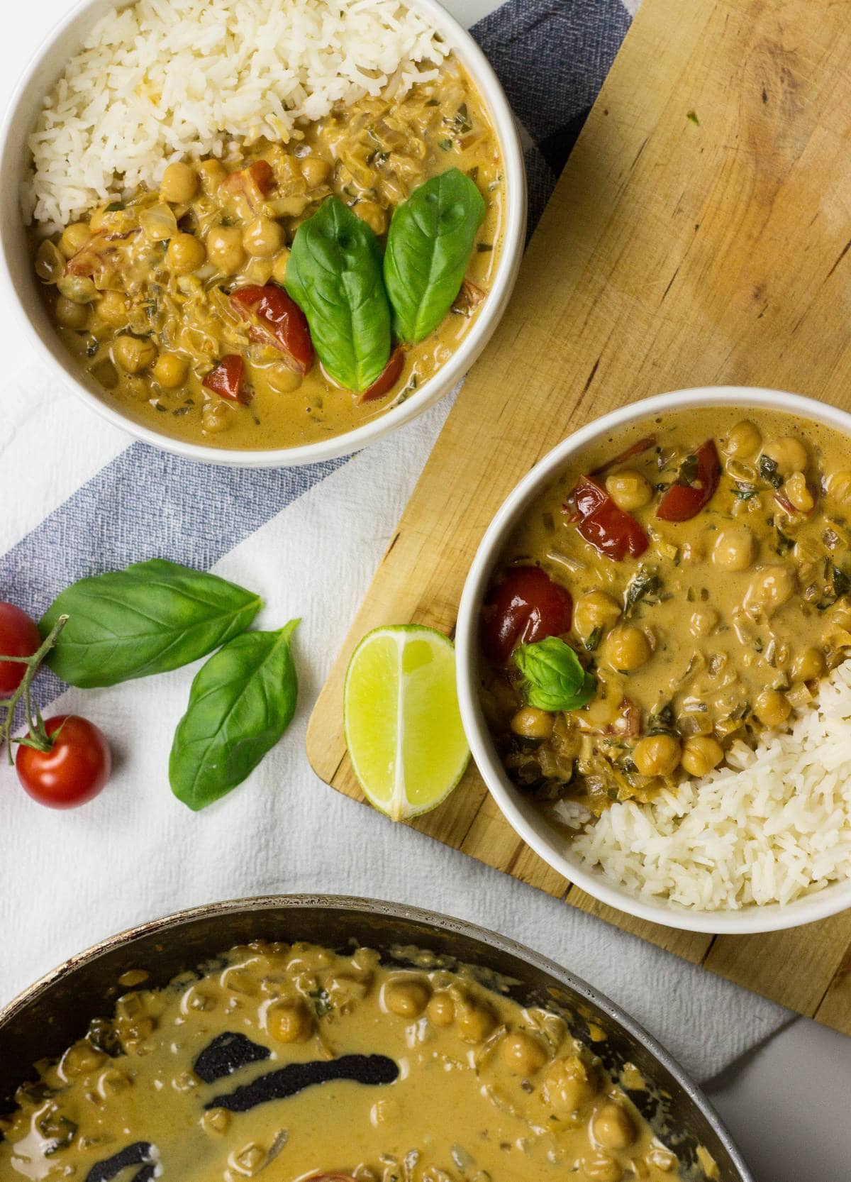 Two bowls with Vegan Chickpea Curry. Next to them, there are some tomatoes, fresh basilic leaves, and a piece of lime | HurryTheFoodUp