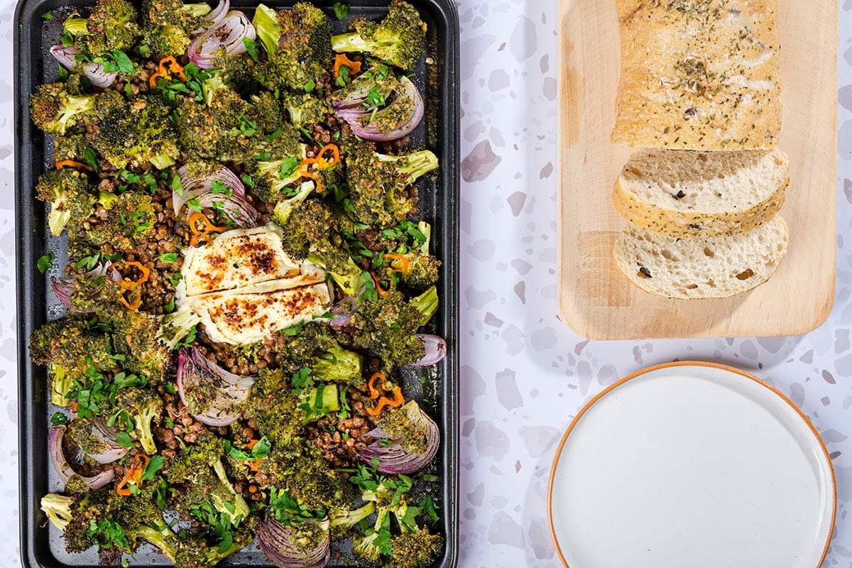 The Roasted Broccoli Halloumi Traybake is on a grey surface next to the wooden board with bread slices and empty white plate | Hurry The Food Up