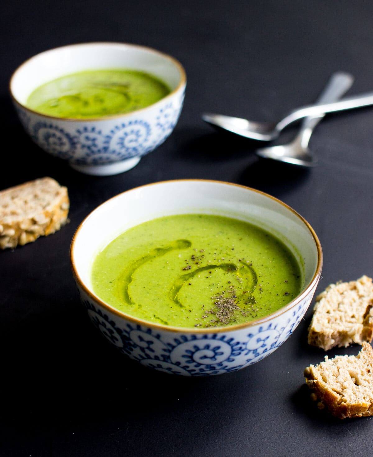 Vertical image of two bowls of Pea and Mint Soup on a black background with spoons and bread | Hurry The Food Up