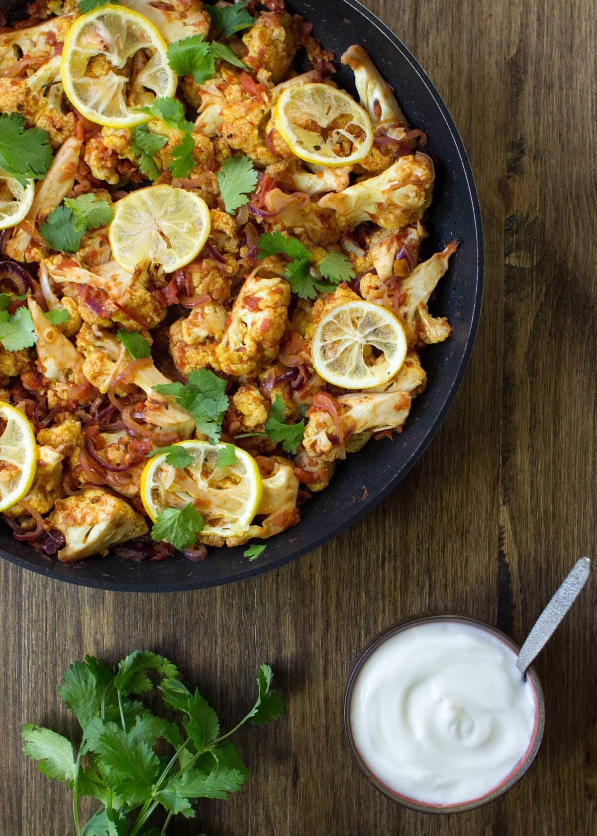 Bird's-eye view of Roasted Cauliflower Curry is in the pan on the wooden surface. Next to it there is a bowl with yogurt and fresh sprig cilantro | Hurry The Food Up