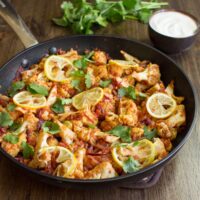 Roasted Cauliflower Curry is in the pan on the wooden surface. Next to it there is a plate with yogurt and fresh sprig cilantro | Hurry The Food Up - 10