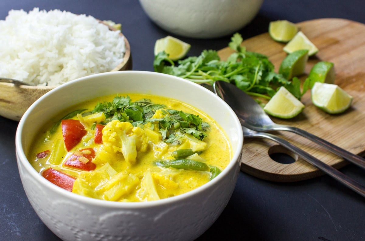 A bowl of Vegan Cauliflower Curry is on the dark-blue surface. Next to it there are a wooden bowl with rice and a wooden board with pieces of lime, a bunch of parsley, and two spoons on it  | Hurry The Food Up