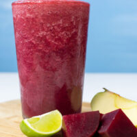 The glass with beetroot smoothie is on the wooden cut board. Near it, there are some pieces of beetroot, lime and apple | Hurry The Food Up - 8
