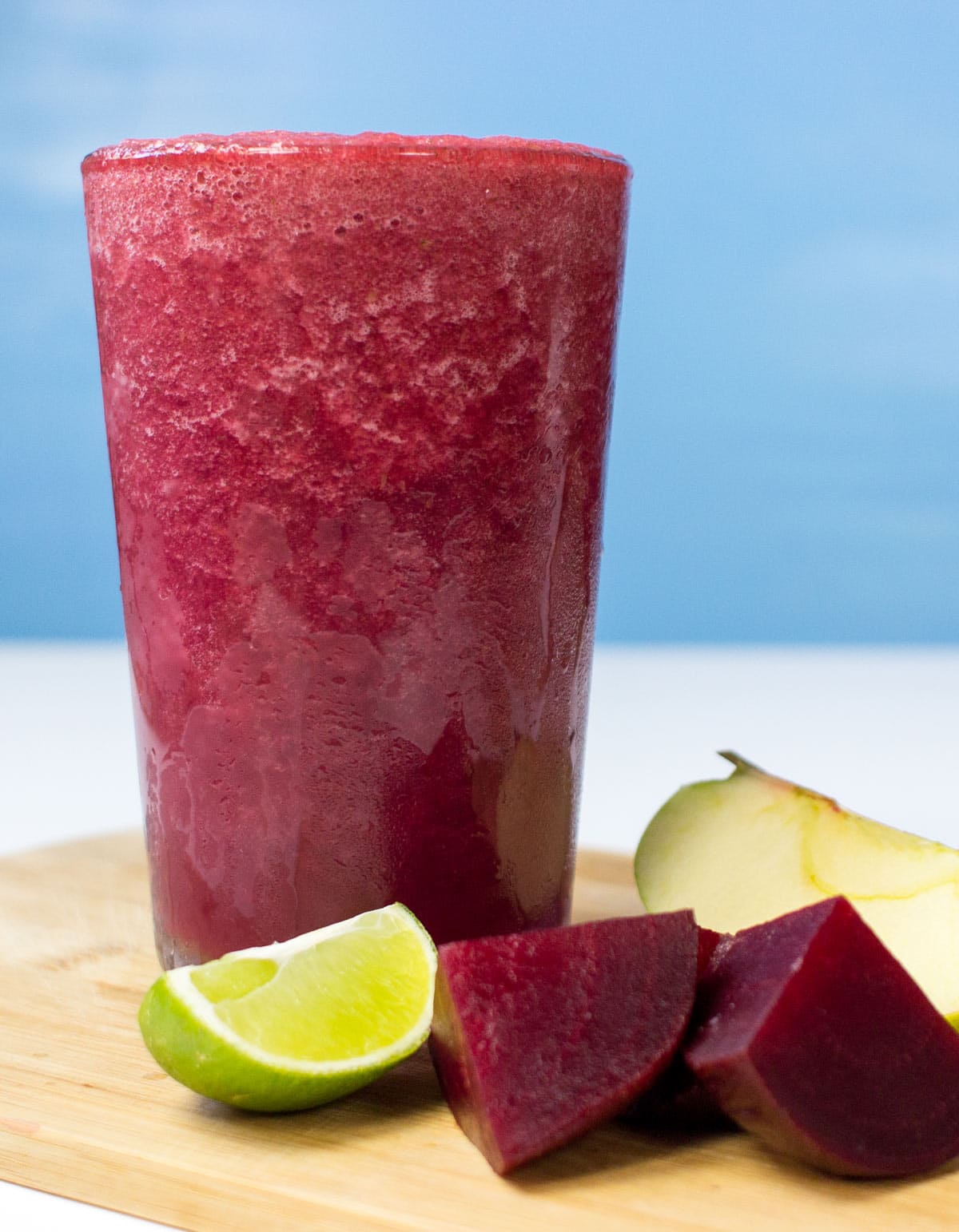 The glass with beetroot smoothie is on the wooden cutting board. There are some pieces of beetroot, lime and apple next to it | Hurry The Food Up