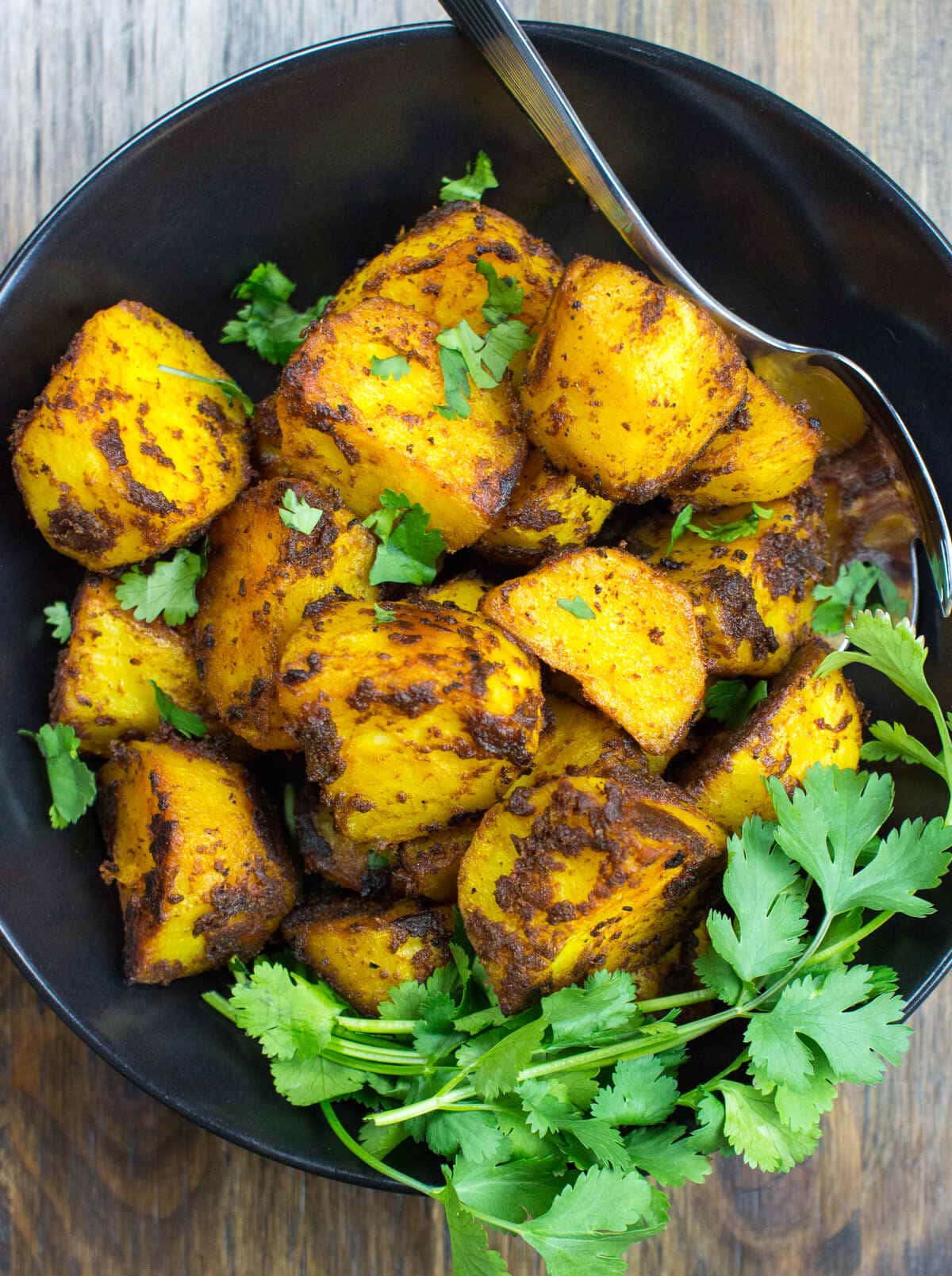 Curried potatoes shot from a bird's eye view, with a spoon and presented with a garnish in a black bowl on a wooden background | Hurry The Food Up