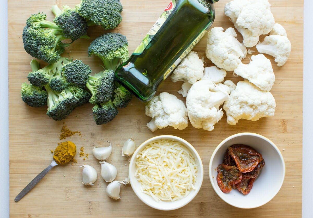 Broccoli, cauliflower florets, a teaspoon of curry, garlic cloves, a bottle of extra virgin olive oil and two small bowls of sundried tomatoes and grated cheese are laid out on a table | Hurry The Food Up