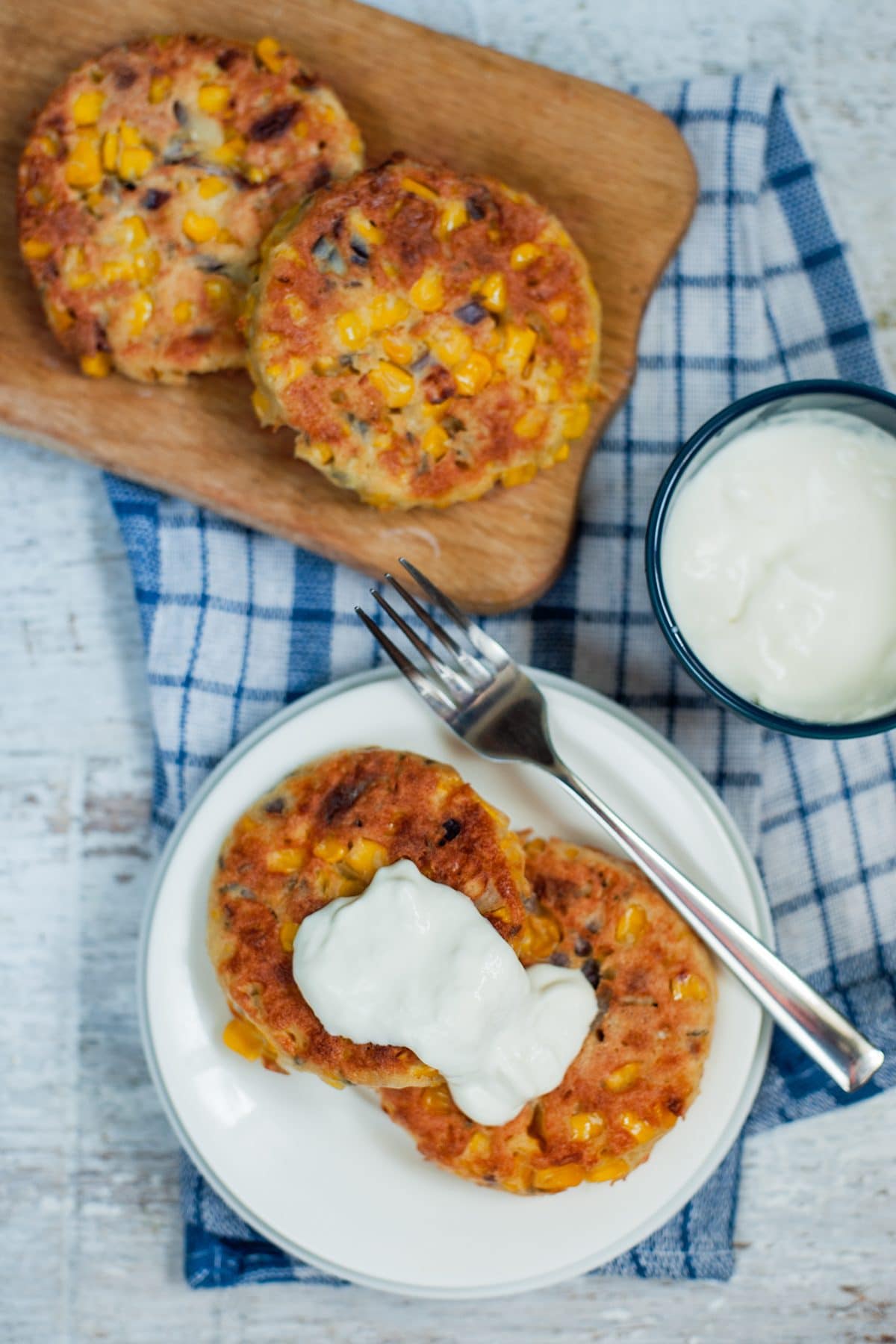 A serving of sweetcorn fritters sits on a white plate with a fork. Next to a board with two fritters on and bowl of sour cream all sat on a blue checked cloth | Hurry The Food Up