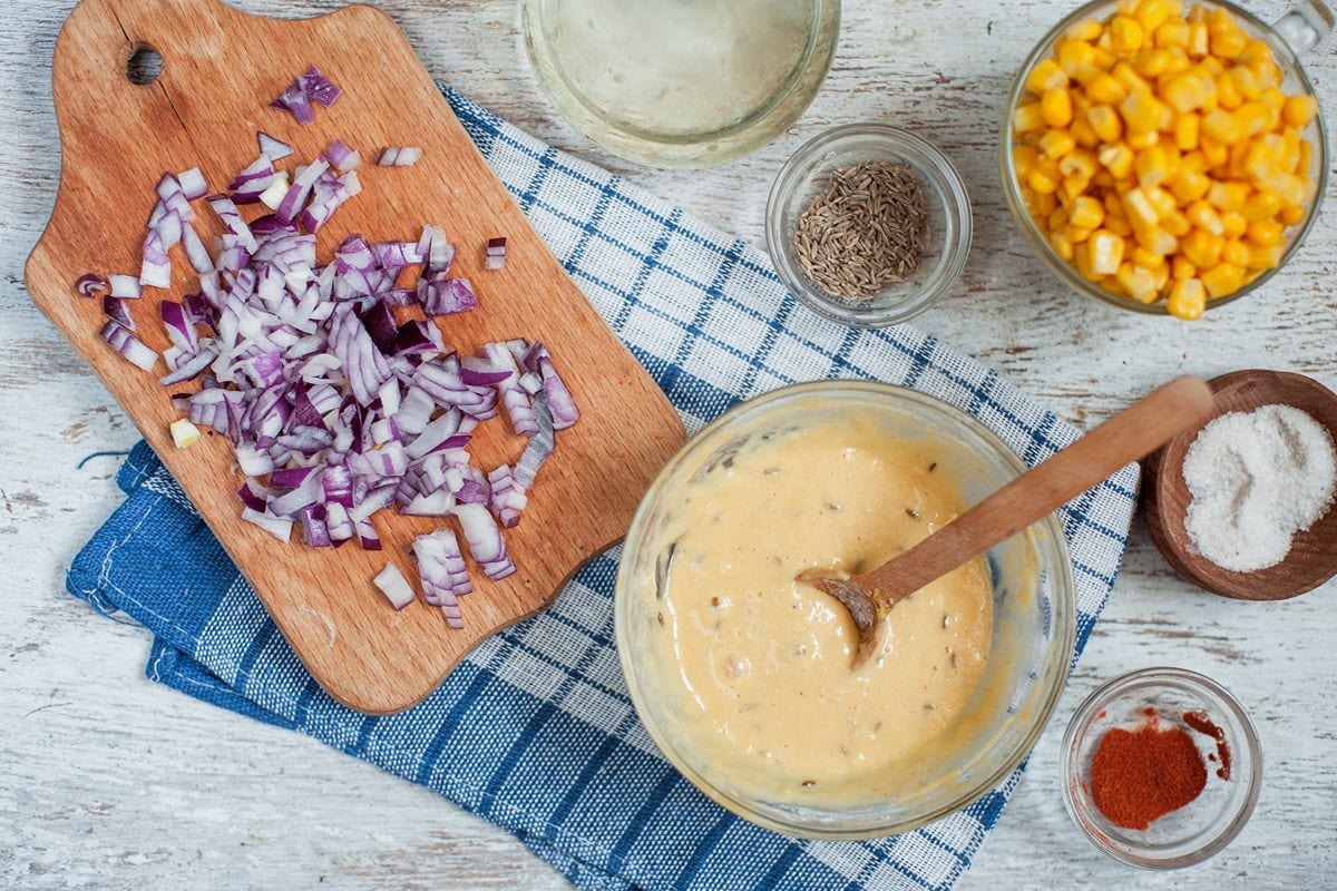 The batter for the fritters is in a glass bowl with a wooden spoon in. Next to it are the other ingredients in little bowls and chopping board with diced red onion on top. | Hurry The Food Up