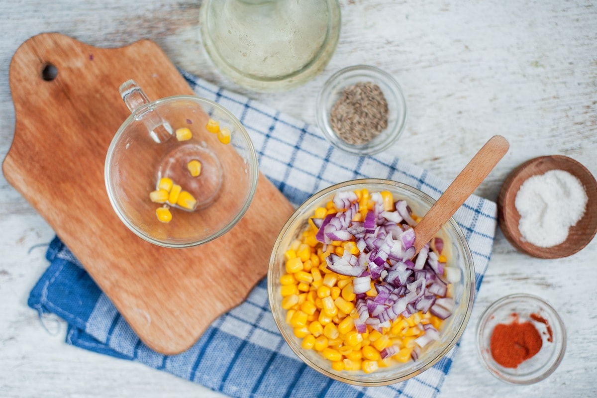 The glass bowl has sweetcorn and red onion in and a wooden spoon for mixing. Around it are small bowls with other ingredients in and a chopping board sat on a blue checked cloth on a wooden table | Hurry The Food Up