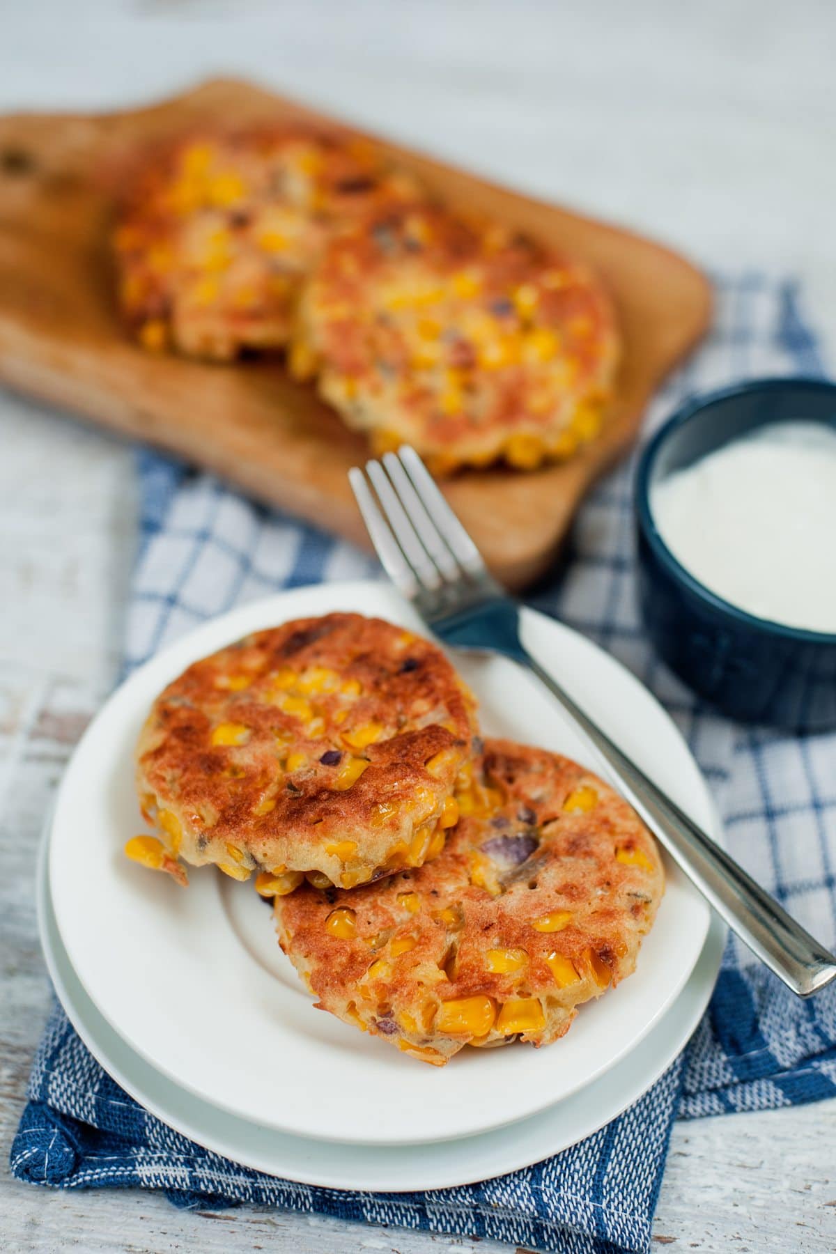 Two sweetcorn fritters and fork are sat on a white plate on top of a blue checked cloth. In the background is a pot of sour cream and two more fritters on a board | Hurry The Food Up