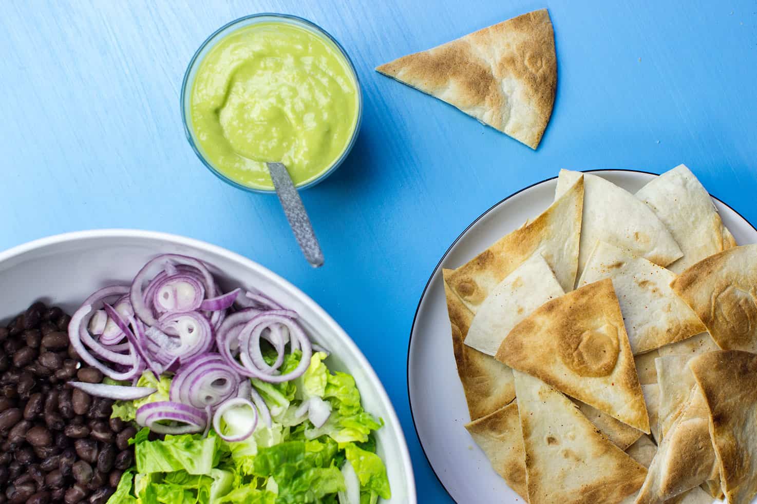 An avocado salad dressing is sat on a table next to a black bean, lettuce and onion salad and a plate of tortilla chips | hurrythefoodup.com