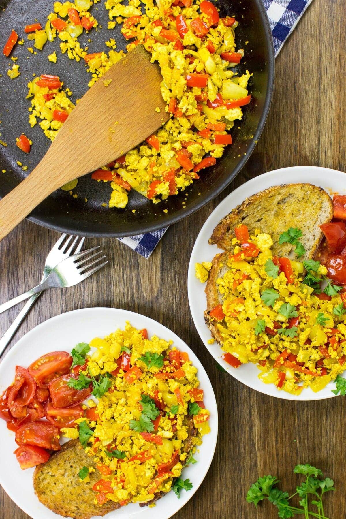 Two plates of scrambled tofu with toast, a frying pan with scrambled tofu and a wooden spatula as well as two forks are placed on a table | Hurry The Food Up