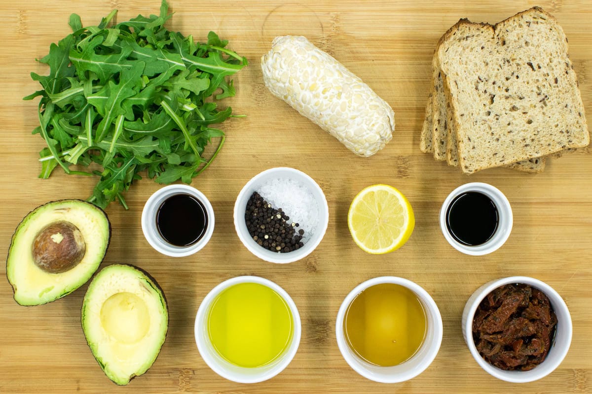 Olive oil, an avocado split in half, salt and pepper, arugula, sundried tomatoes, maple syrup, half a lemon, bread slices, soy sauce and tempeh are placed in small white bowls over a table | Hurry The Food Up