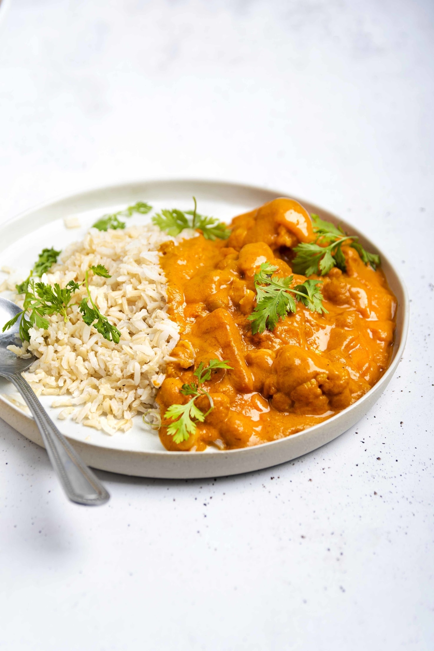 A plate of vegan tikka masala on a white surface, served with brown rice and garnished with parsley, a spoon to eat it with on the same plate | Hurry The Food Up