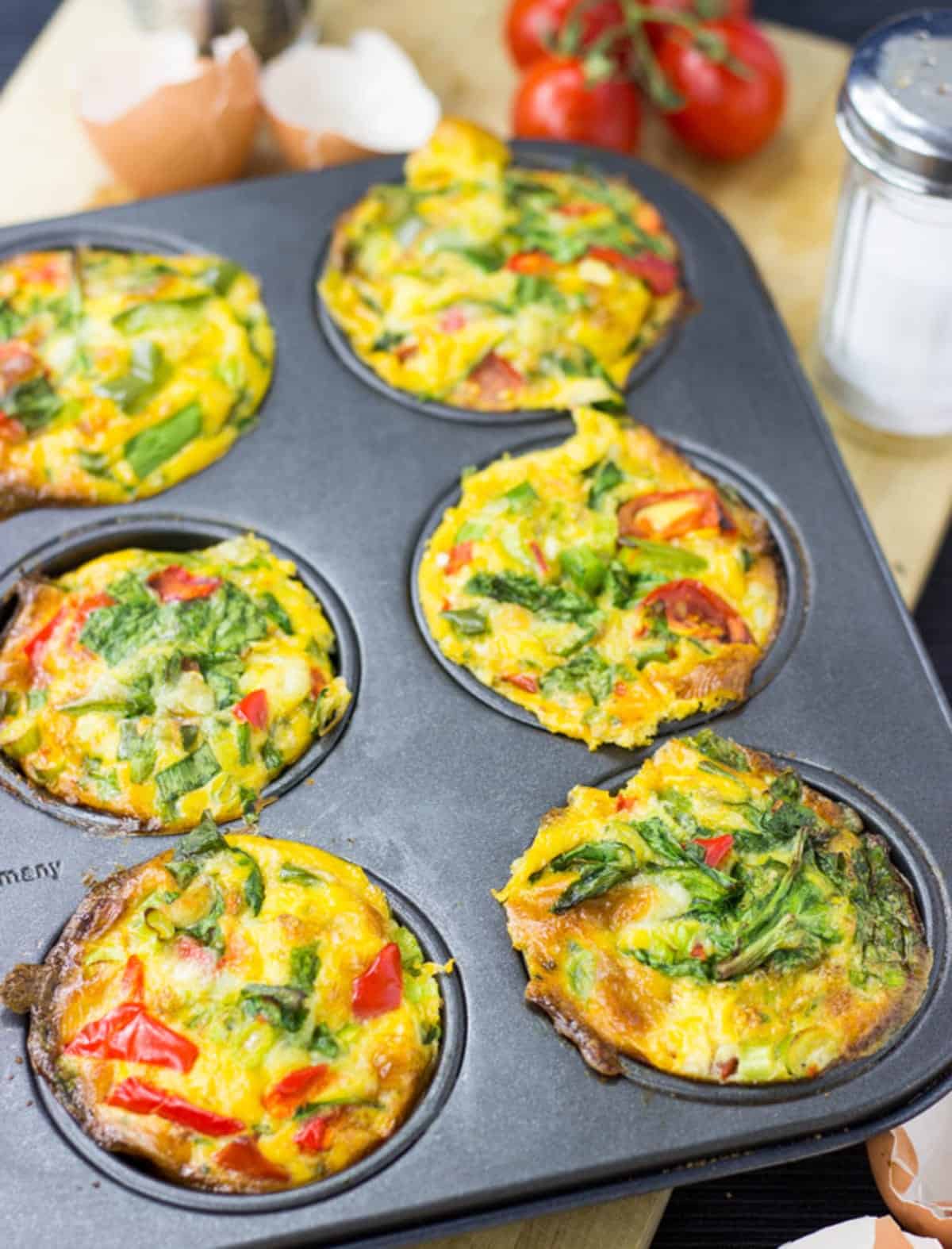 A muffin tray full of cooked breakfast egg muffins . Some ingredients are visible in the background. | Hurry The Food Up