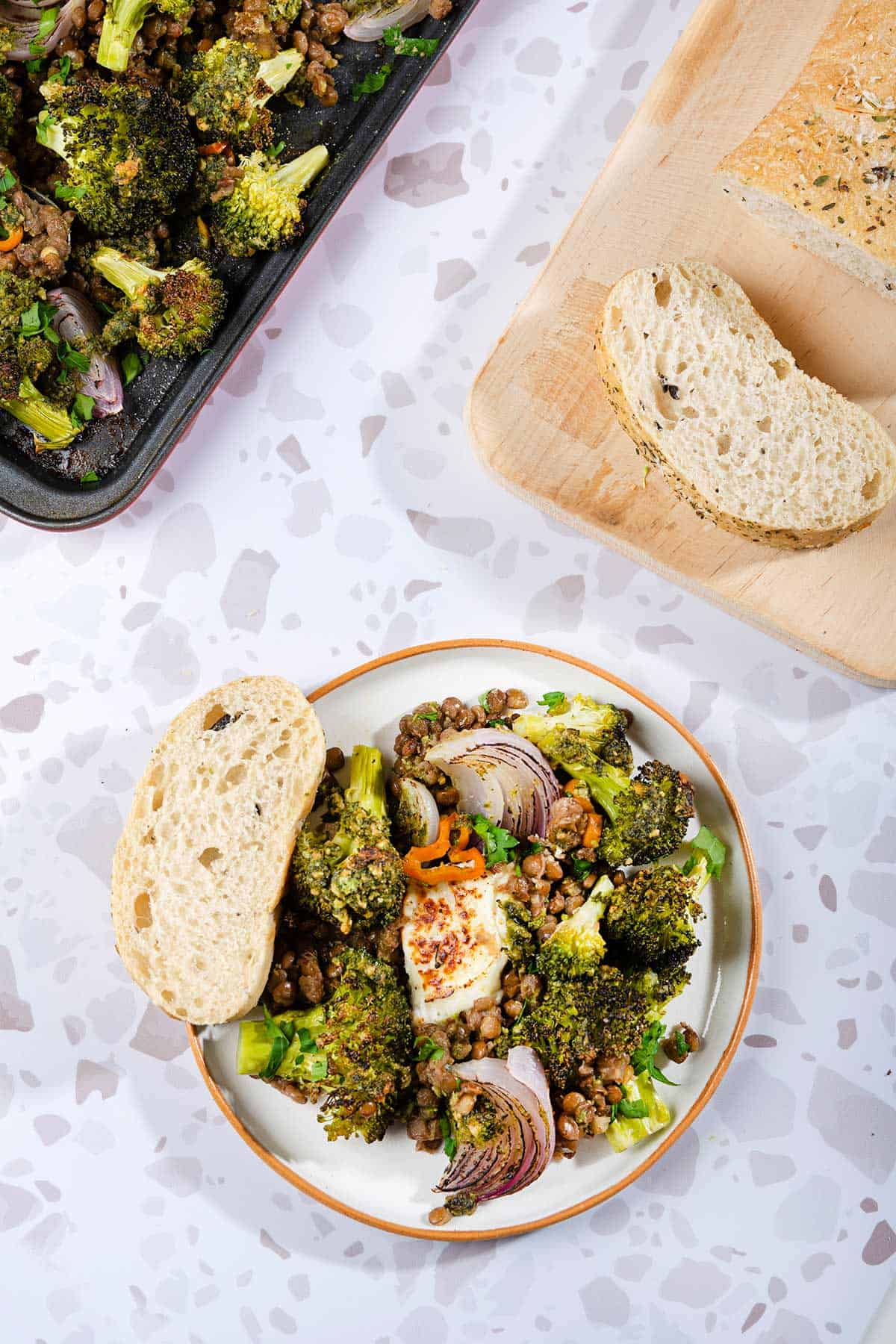 A birds eye view of a Roasted Broccoli Halloumi that is served on a white plate next to the baking tray and wooden board with bread slices | Hurry The Food Up