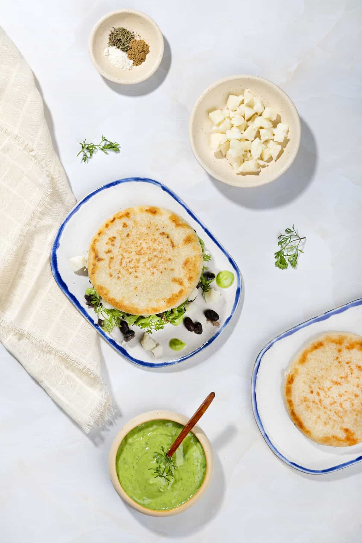 Arepas ingredients like cornmeal, vegetable broth, onion powder, white onion, black beans, avocado, parsley etc. | Hurry The Food Up A birds eye view of plated aripas with black beans, with a bowl of green sauce, a bowl of crumbled feta, a plate of unfilled arepas, spices | Hurry The Food Up