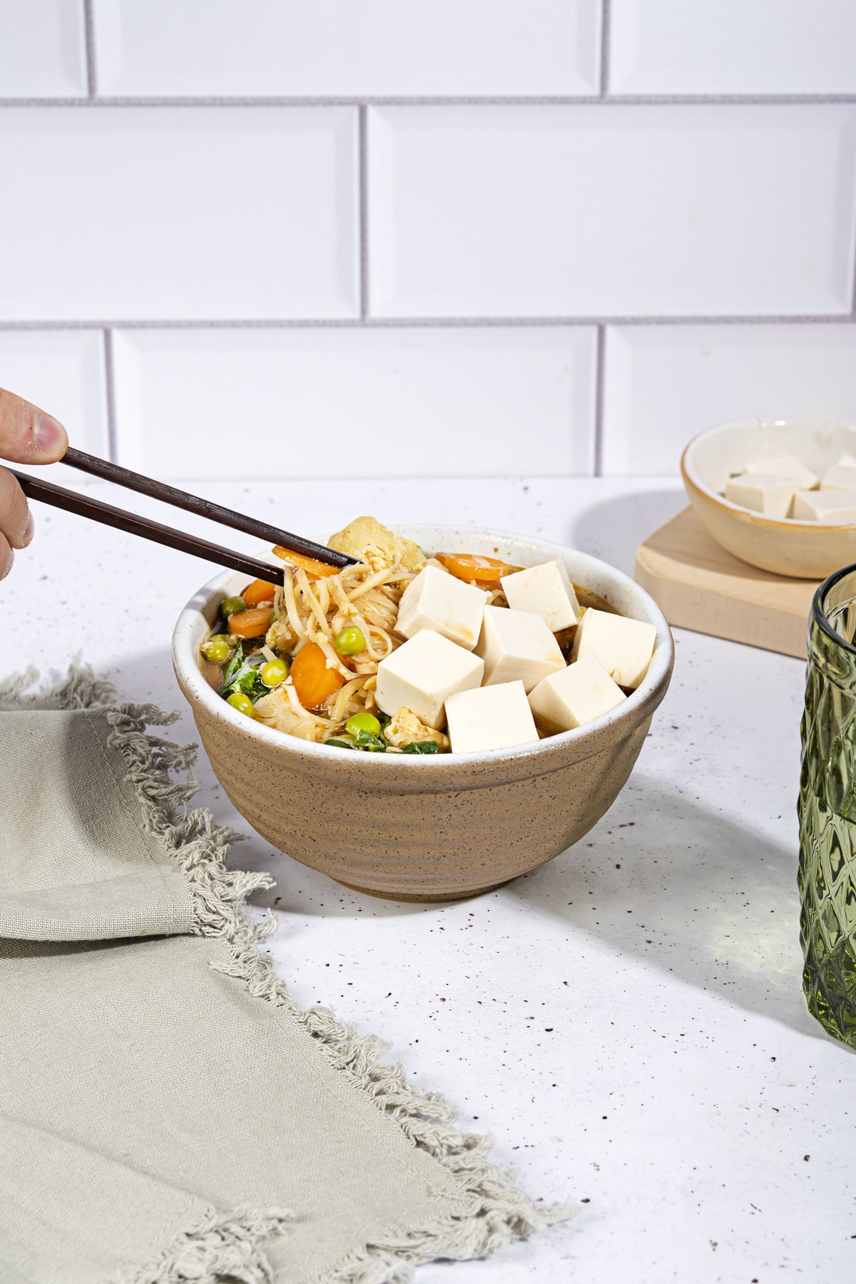 A person is picking up noodles from the bowl of egg drop soup that is on the white table along the bowl with tofu cubes, grey towel and a glass of water | Hurry The Food Up