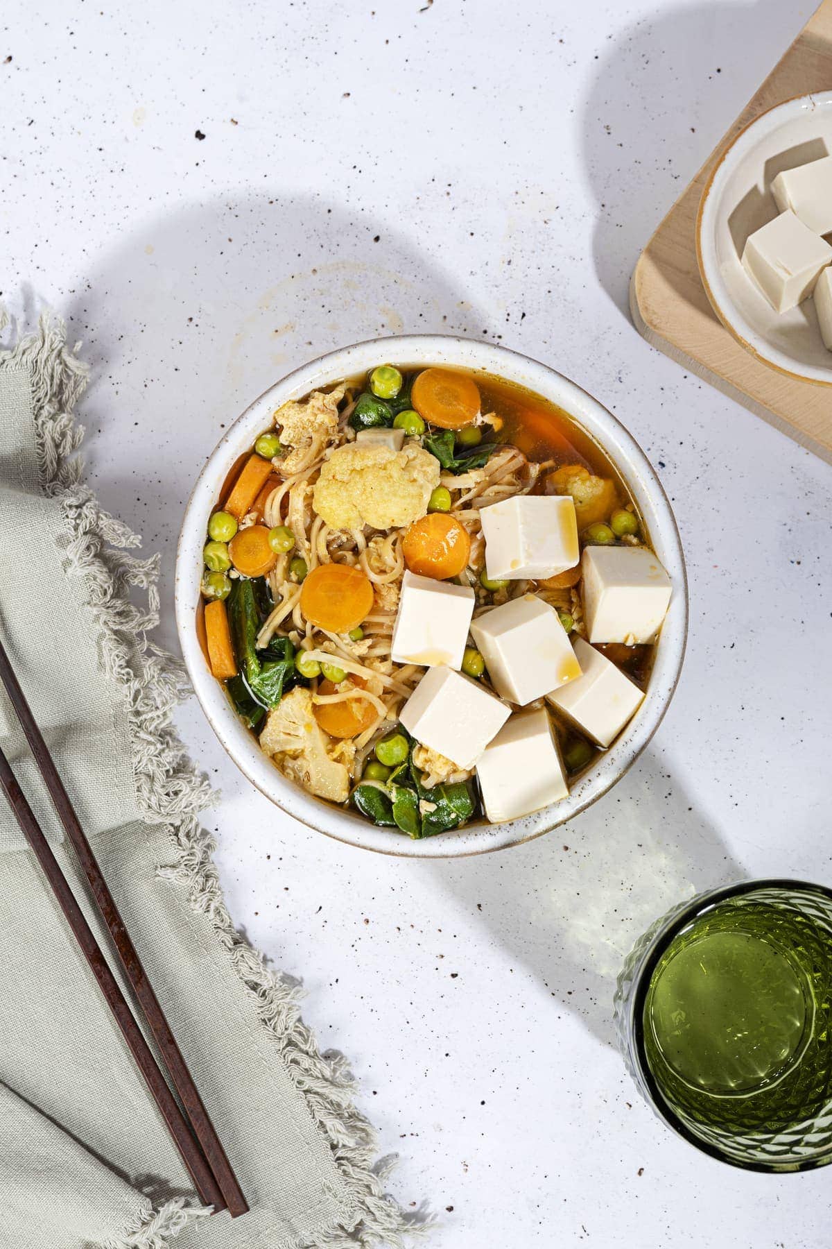 A bird eye view of the bowl with egg drop soup is on the white table along the bowl with tofu cubes, black chopsticks on a grey towel and a glass of water | Hurry The Food Up