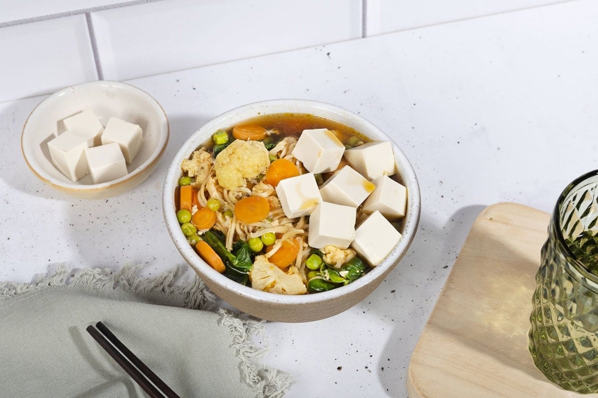 The bowl with egg drop soup is on the white table along the bowl with tofu cubes, black chopsticks and a glass of water that is on the chopping board | Hurry The Food Up