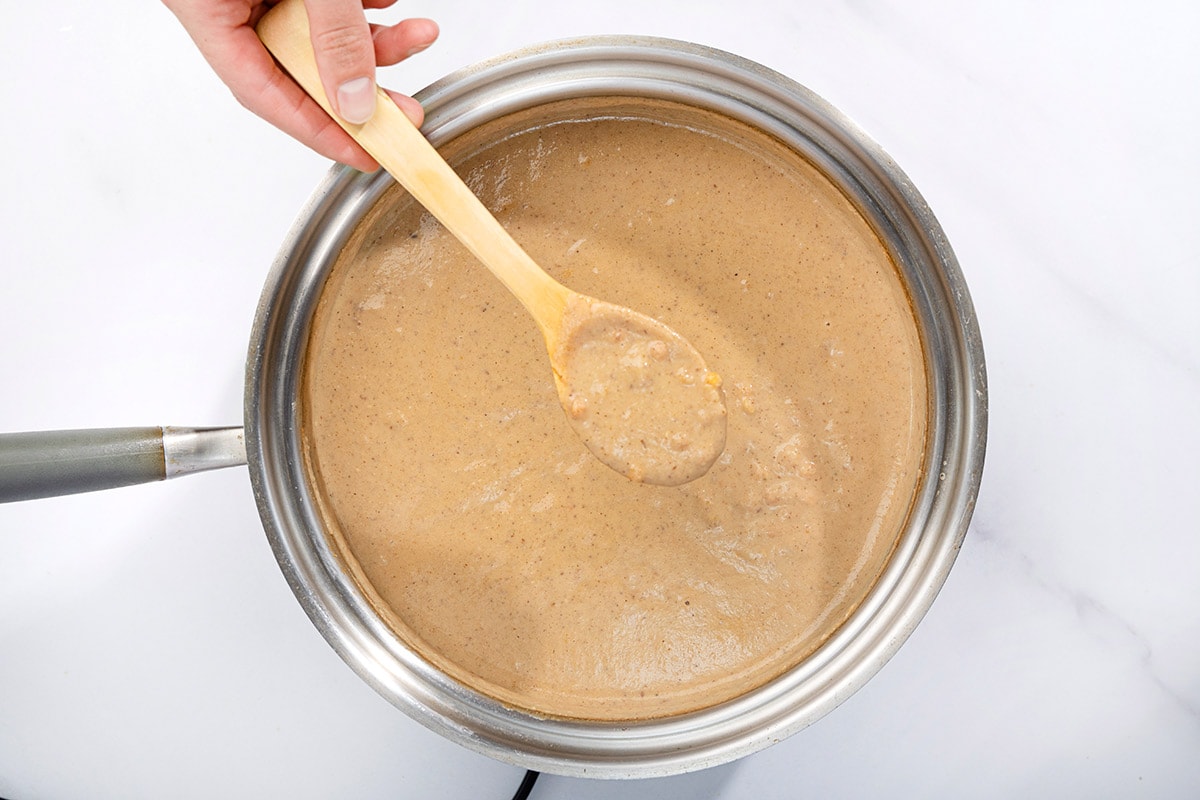 On a marbled surface sits a saucepan full of vegan cauliflower soup. A person is holding a spoon over the saucepan with some soup on it | Hurry The Food Up