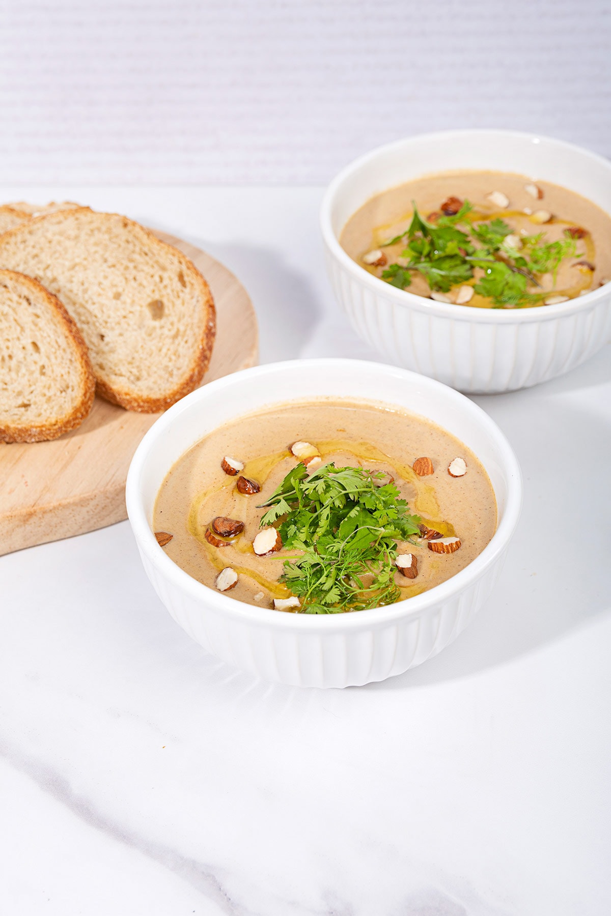 On a marbled table sits two white bowls of creamy vegan cauliflower soup, topped with olive oil, sliced almonds and cilantro. Next to them is a board with some slices of bread | Hurry The Food Up