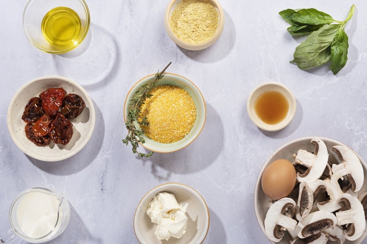 Ingredients sit in different sized bowls on a marbled surface. There is olive oil, sun-dried tomatoes, a glass jug of milk, nutritional yeast, uncooked polenta, a sprig of thyme, some mushrooms, cream cheese, basil, vinegar and an egg. They all sit on a marbled surface | Hurry The Food Up