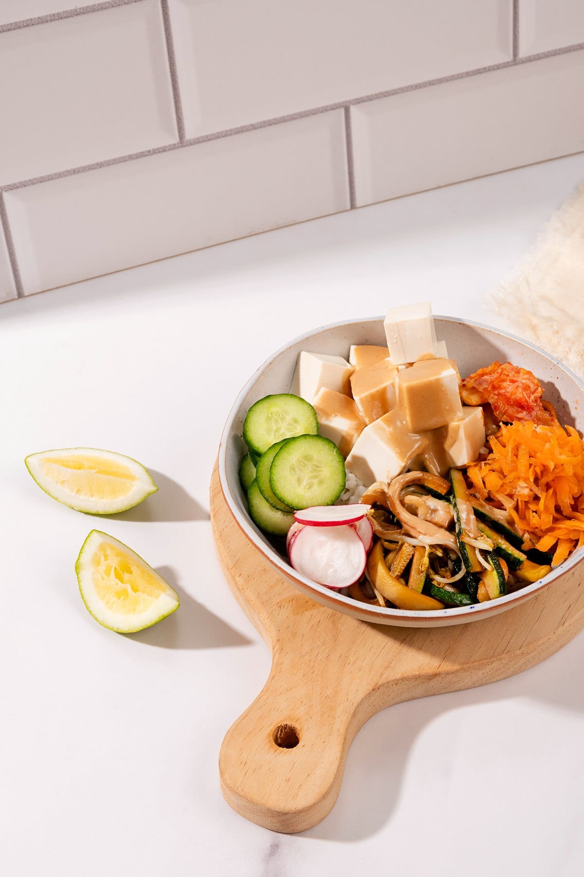 A portrait image of a bibimbap bowl sitting on a chopping board on a white table. Next to the bowl are two lemon wedges and a cream cloth | Hurry The Food Up