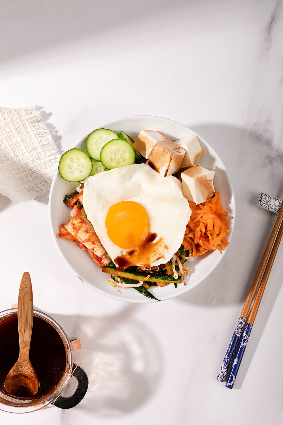 A birds-eye view of a vegetarian bibimbap in a large bowl. Underneath is a pot of soy sauce and a wooden spoon, to the right is a pair of chop sticks | Hurry The Food Up