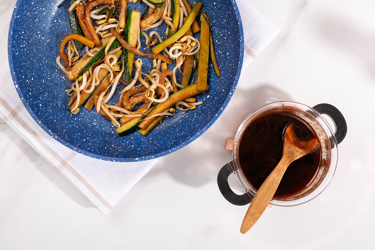 A birds-eye view of a blue frying pan holding zucchini and bean sprouts on a white check tea towel on a white table. Next to the pan is a pot of soy sauce with a wooden spoon in it | Hurry The Food Up