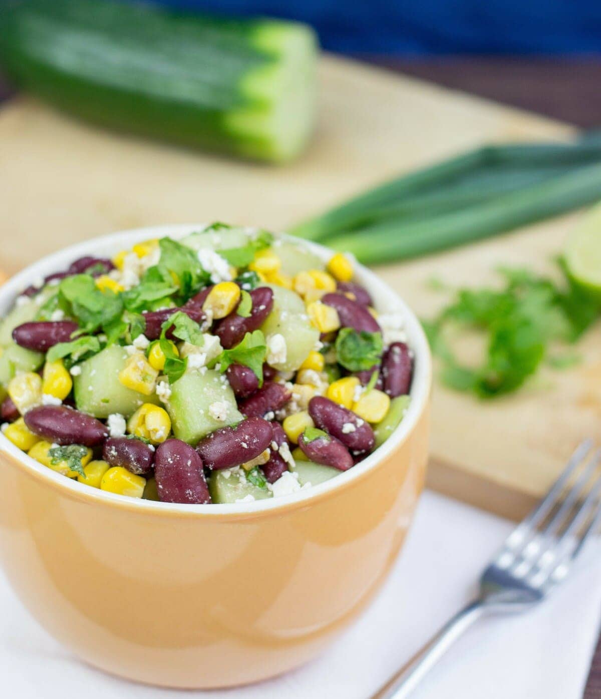 High-protein kidney bean salad is plated in a ceramic bowl and placed on a white napkin with a fork. Spring onions, a cucumber and parsley leaves are in the distance | Hurry The Food Up