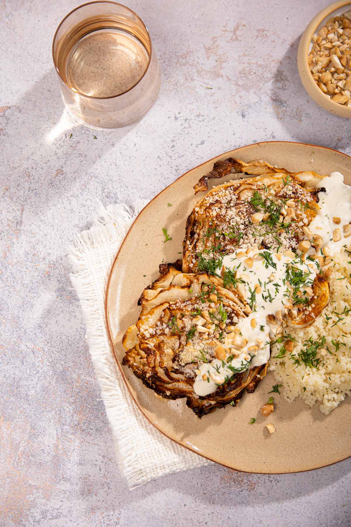 Roasted cabbage and couscous on a ceramic plate, sitting on a cream cloth on a stone table. Above is a glass of water and bowl of nuts | Hurry The Food Up