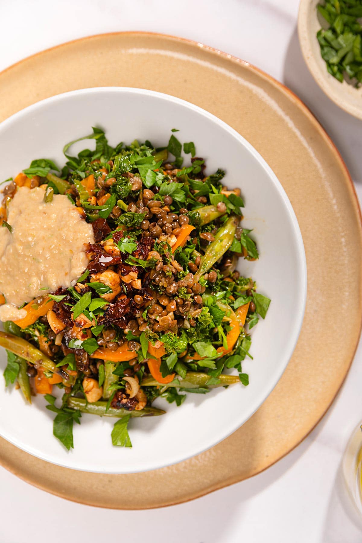 Overview of lentils with vegetables that are served in the white plate that stands on the beige plate which is on the white surface. Next to it there is a small plate with chopped parsley | Hurry The Food Up