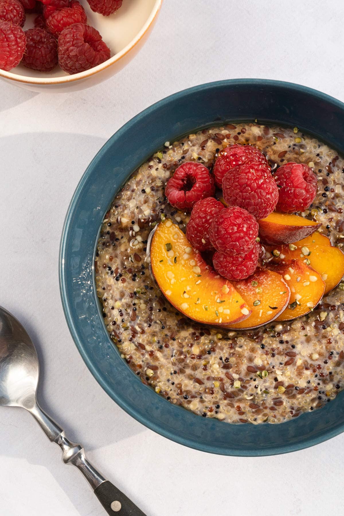 Bird's-eye view of fruity quinoa porridge that is served in the bowl with pieces of peach and raspberries. There are a bowl with raspberries and a spoon next to it | Hurry The Food Up