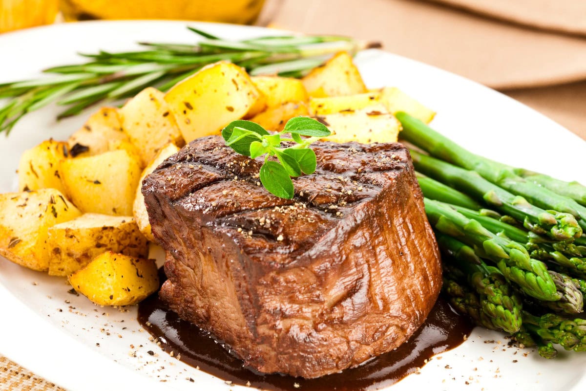 A juicy piece of filet mignon lies on the white plate with potatoes and asparagus.Behind it, there is a branch of rosemary | Hurry The Food Up