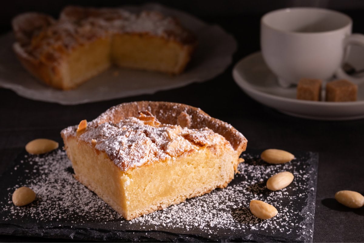 A piece of frangipane lies on the dark surface decorated with some peanuts and sugar powder. Behind it, there is a cup and frangipane | Hurry The Food Up