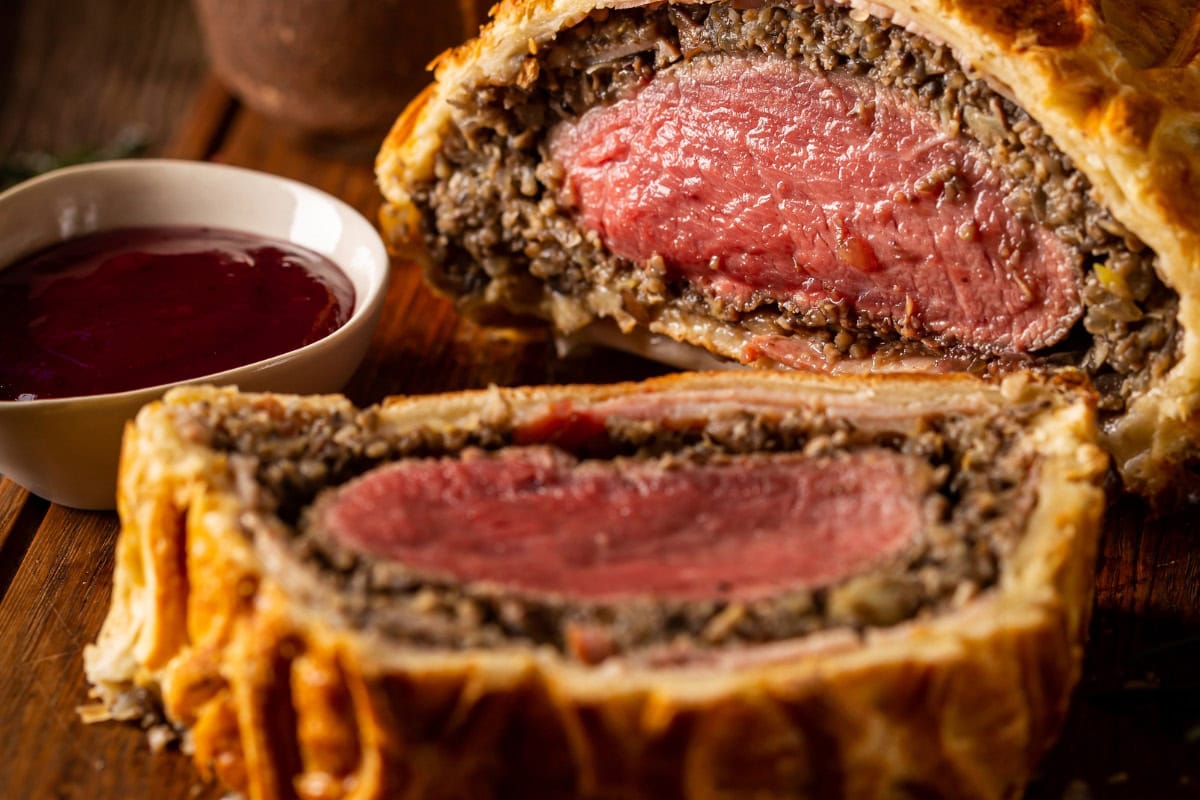 Juicy beef wellington lies on the wooden surface. Next to it, there is small white bowl with souce | Hurry The Food Up