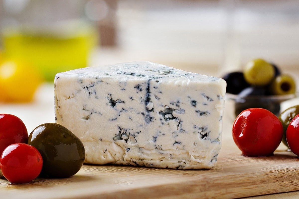 A piece of blue cheese is on the wooden board. Next to it, there are some olives | Hurry The Food Up